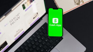 Smartphone showing Cash App screen on laptop keyboard, next to glasses and notebook.