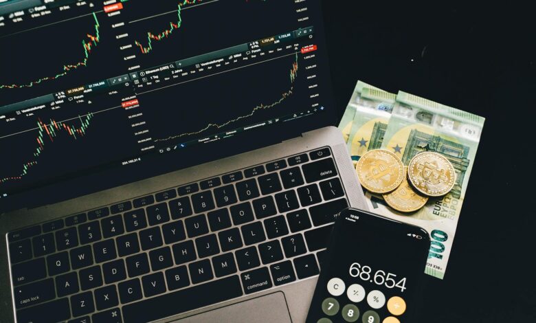 A laptop displaying stock charts with Bitcoin, Euros, and a cellphone calculator, showcasing financial analysis.